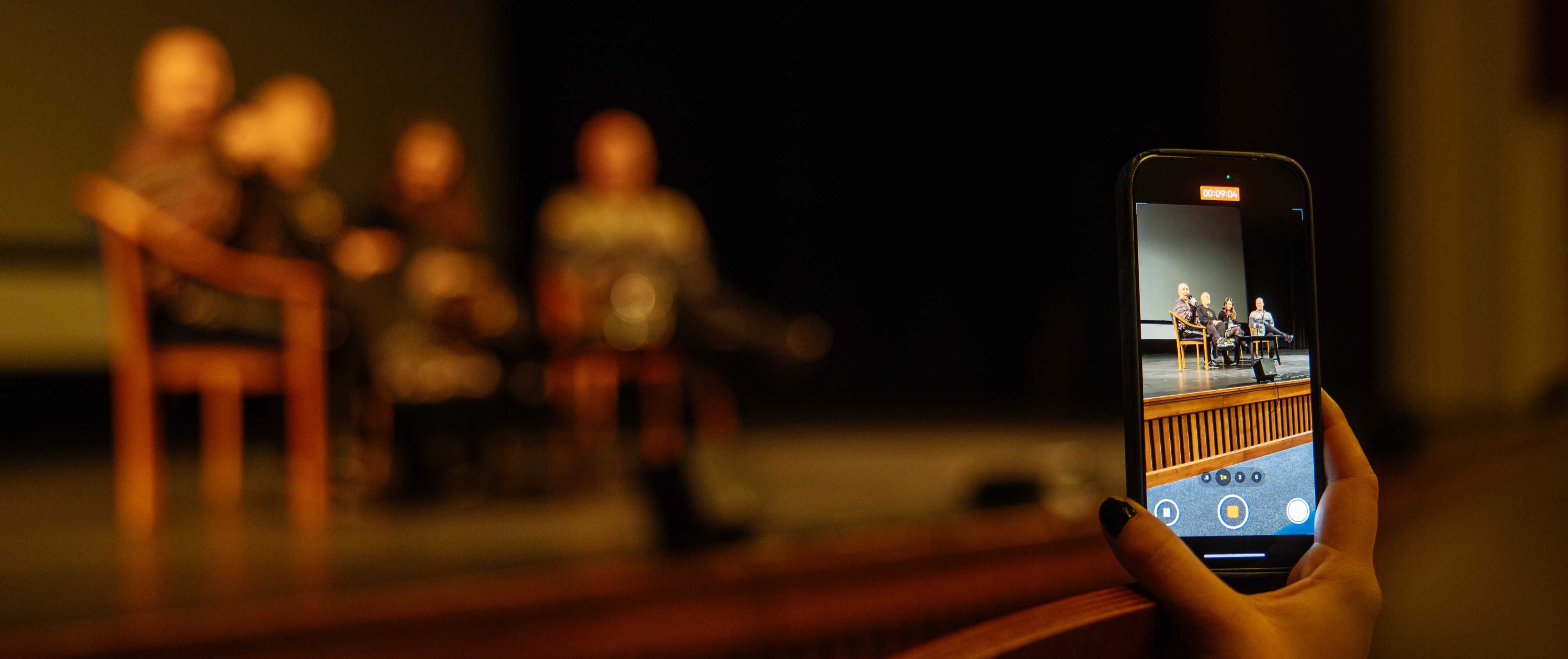 Ruka držící mobilní telefon zachycuje živý záznam diskuse na pódiu. Zatímco skutečný obraz účastníků debaty je rozostřený, obrazovka telefonu je ostrá a jasně ukazuje debatující sedící na jevišti.