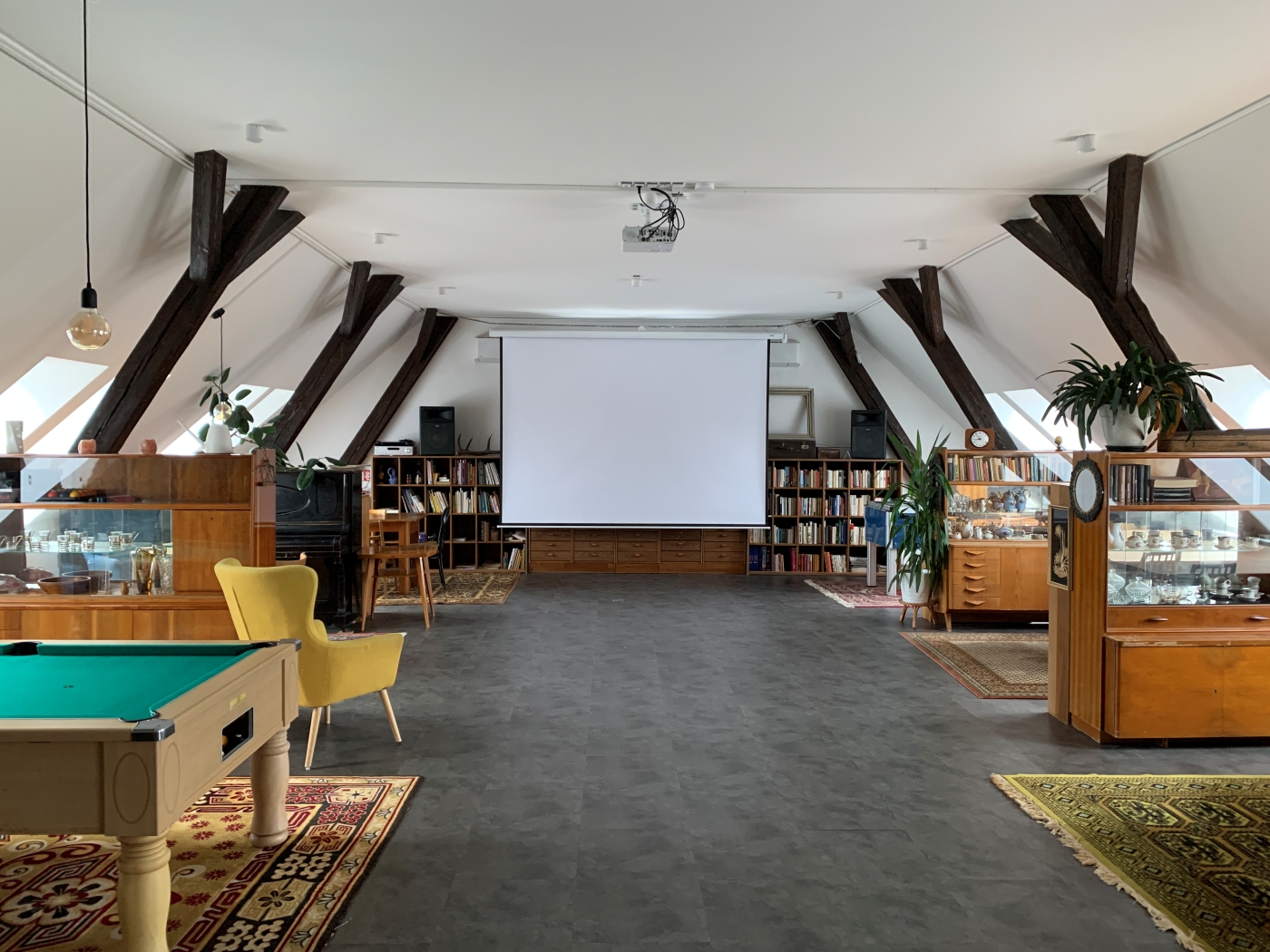 Prostorný podkrovní interiér s dřevěnými trámy, knihovnami po stranách a promítacím plátnem uprostřed.