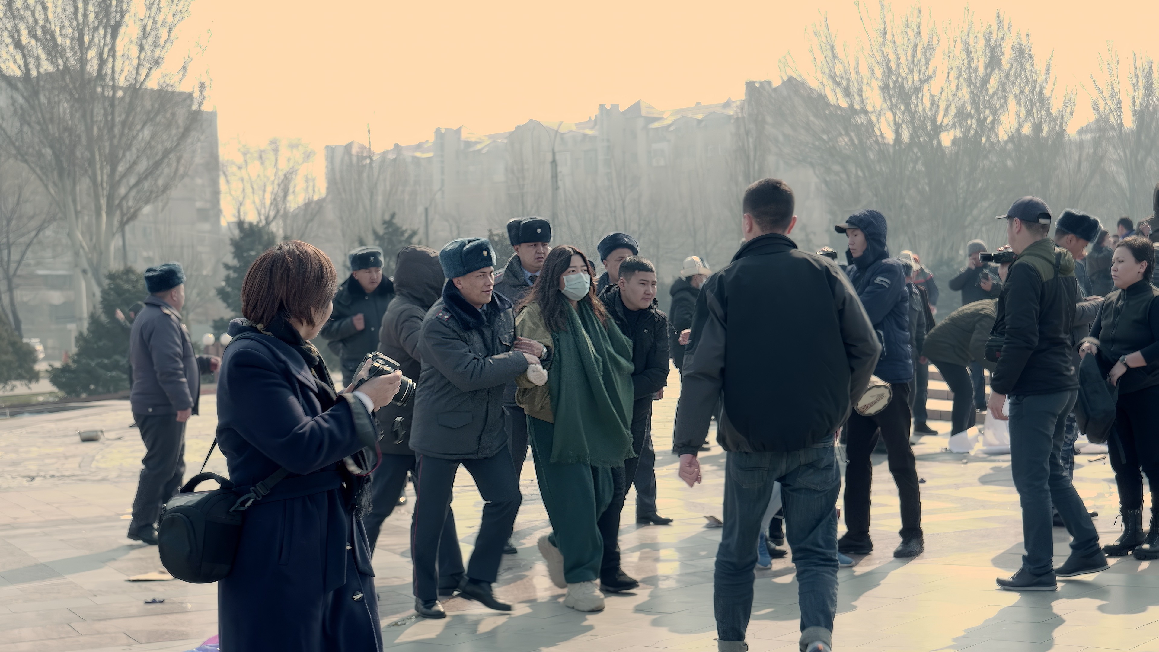 Skupina lidí venku na náměstí, někteří fotografují, jiní se procházejí, dva muži vedou ženu v roušce.