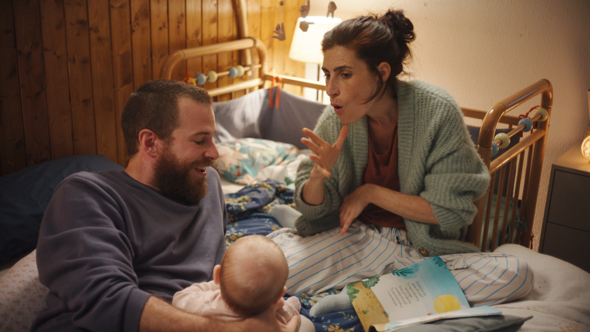 Rodiče sedí na posteli s malým dítětem. Žena na dítě mluví znakovým jazkem. Na posteli leží otevřená kniha.