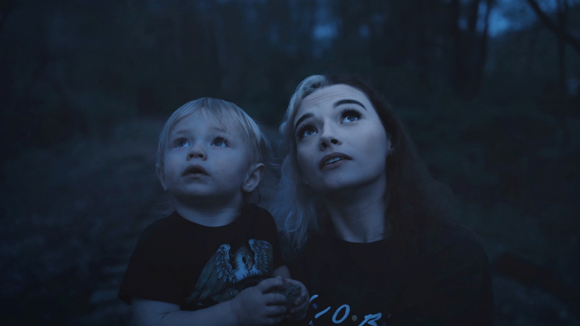 Dospělá osoba a malé dítě v šeru lesa společně vzhlížejí vzhůru, nasvícení jemným modrým světlem.