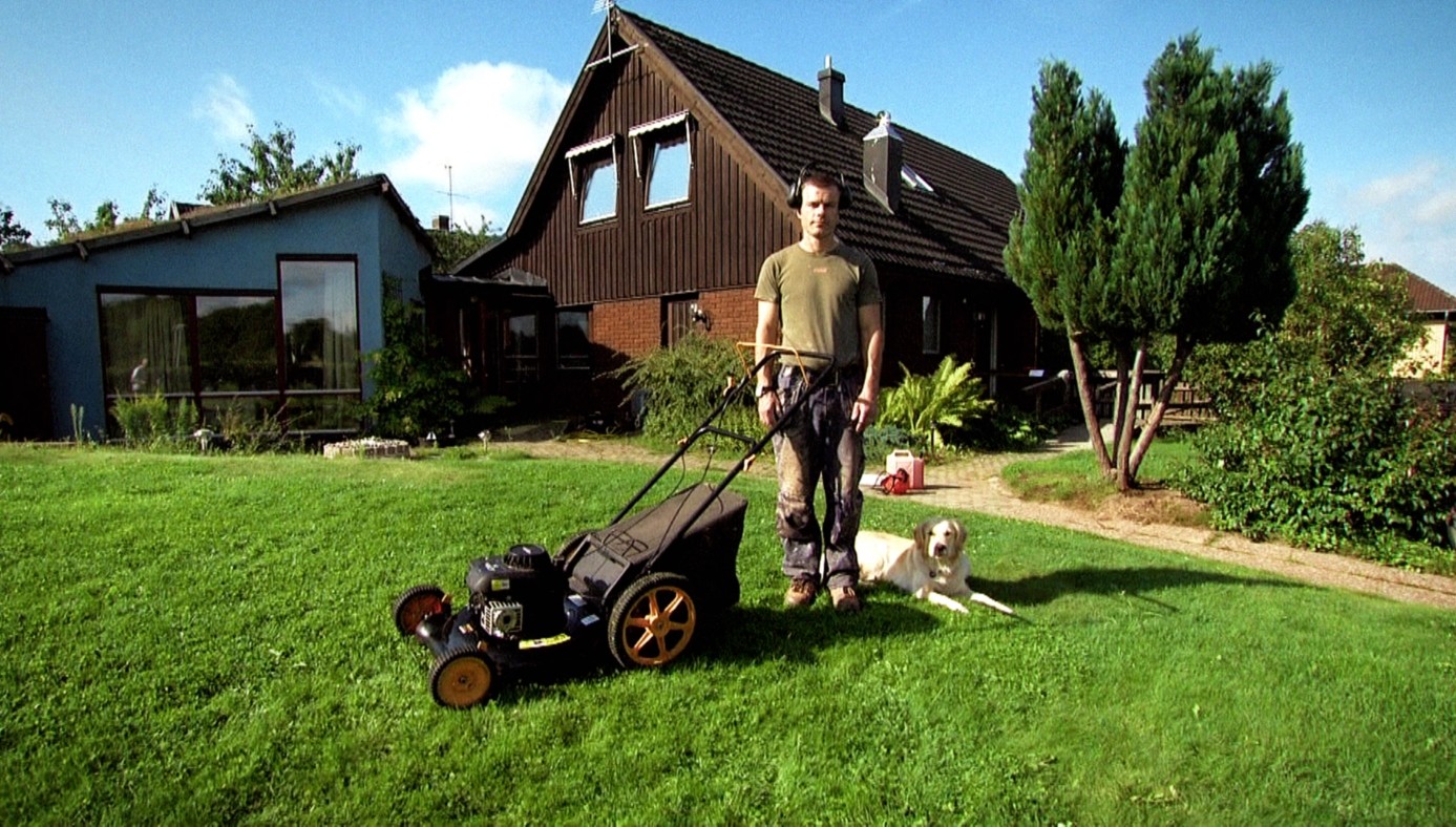 Muž stojí na zahradě u sekačky na trávu, vedle něj leží pes a v pozadí jsou rodinné domy.