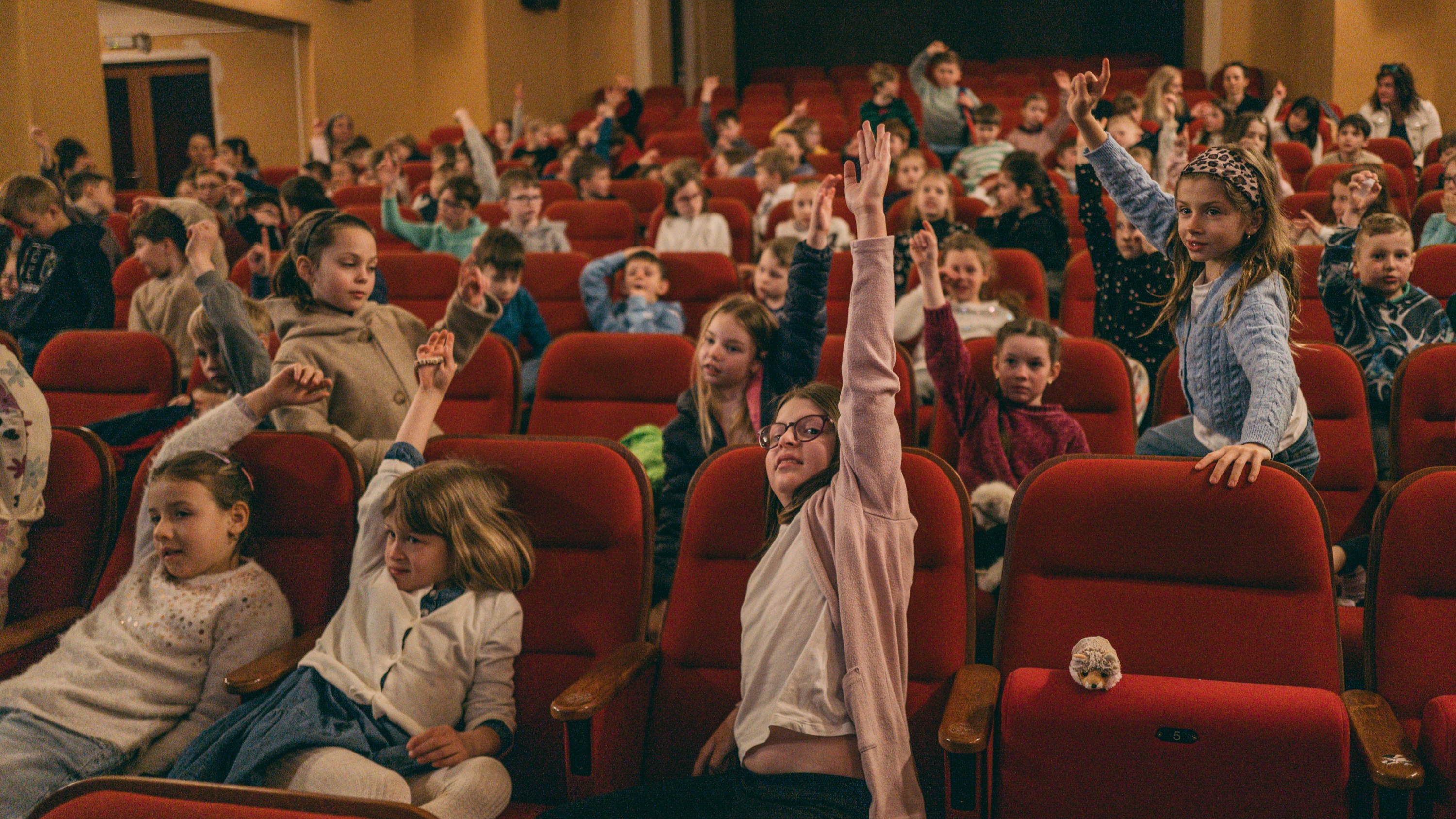 Děti sedí v hledišti kina a aktivně se hlásí, aby odpověděly na dotaz během debaty.