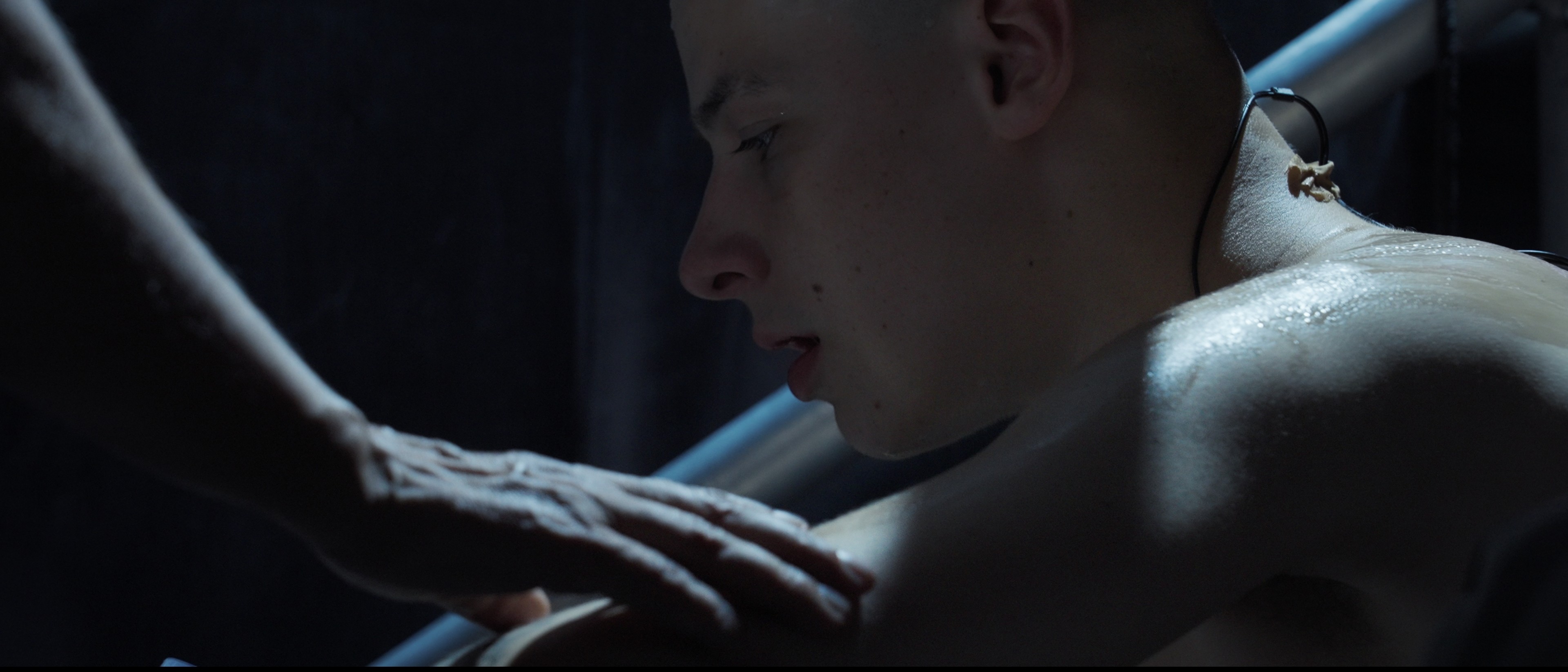 Mladý muž sedí ve tmavém prostředí a někdo mu pokládá ruku na rameno.