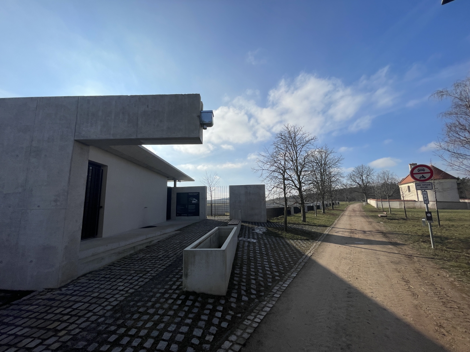 Moderní betonová minimalistická budova stojí na kraji louky. Před ní je dlažba s květináčem, prašná cesta, holé stromy, pole a v dálce stavba s červenou střechou.