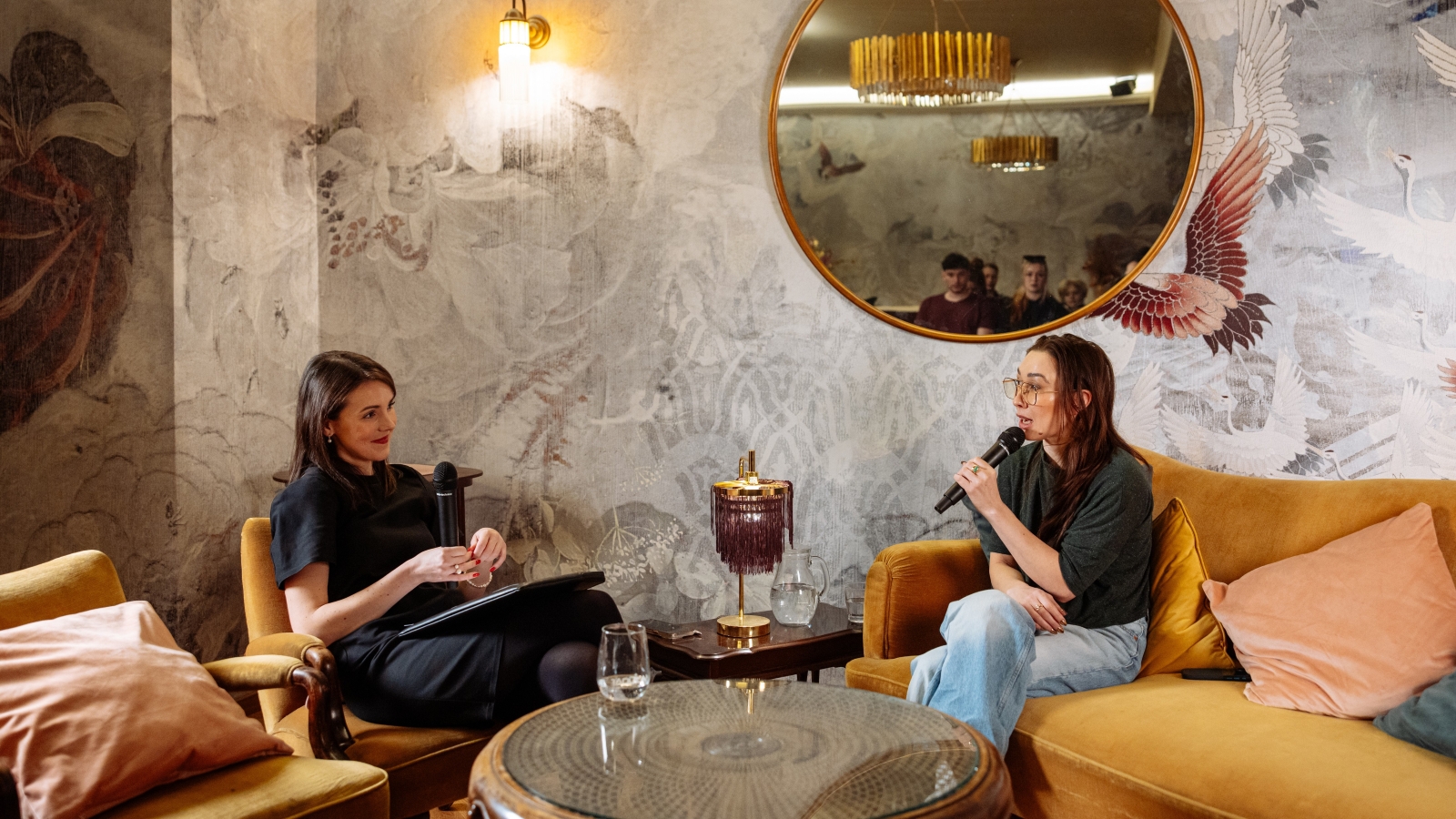 Dvě ženy sedí naproti sobe, jedna na pohovce a druhá v křesle a povídají si.