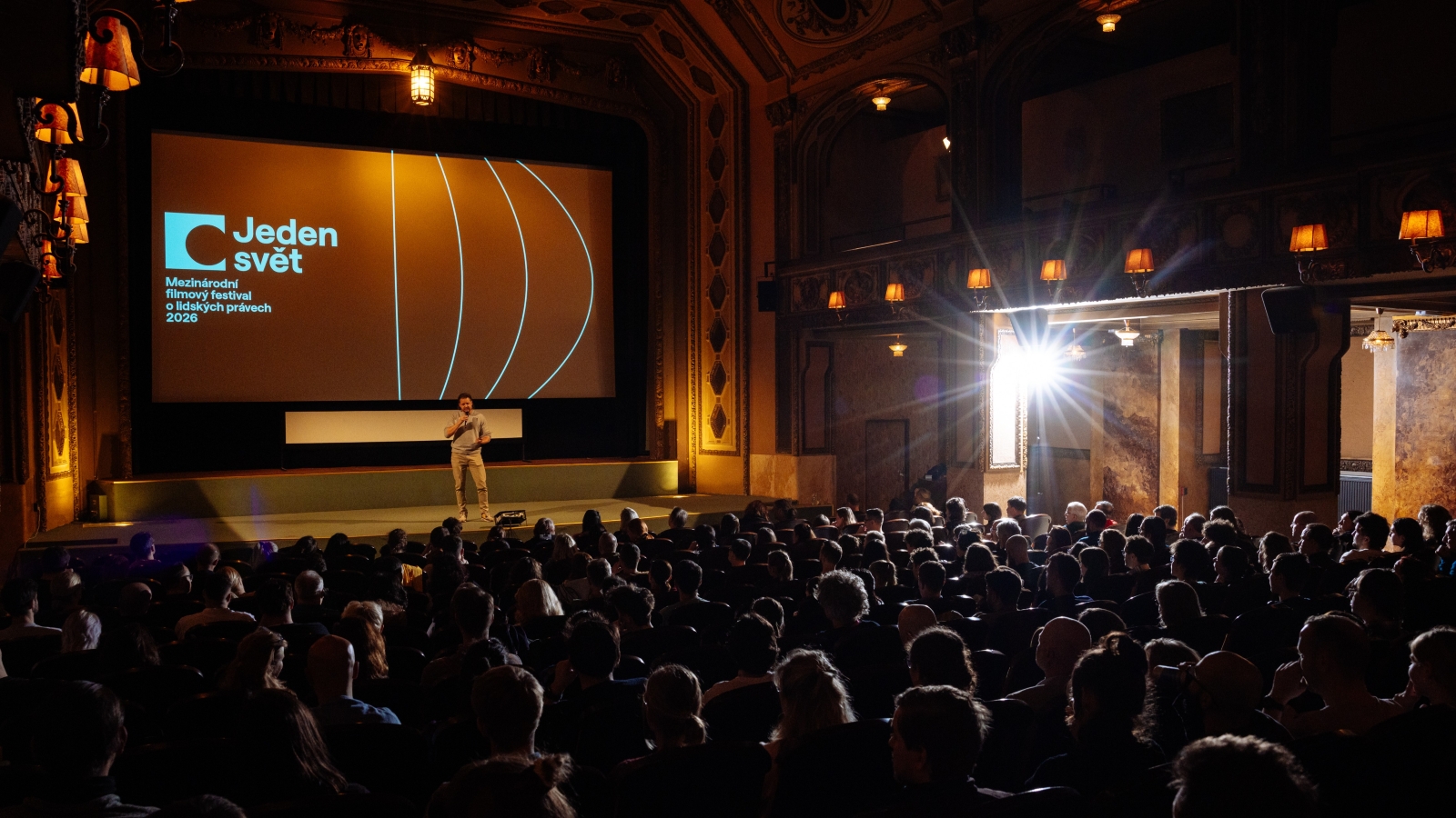 Moderátor stojí na pódiu v historickém kině a hovoří k plnému sálu diváků. Na velkém plátně za ním je promítán vizuál festivalu Jeden svět – Mezinárodní filmový festival o lidských právech 2026.