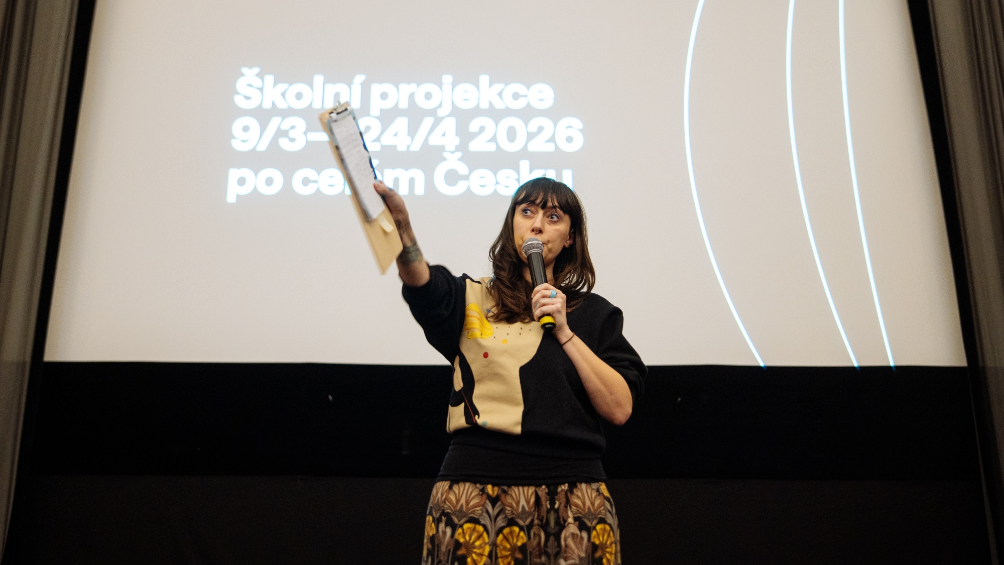 Žena stojí před plátnem, na kterém je napsáno Školní projekce 9/3 - 24/4 2026 po celém Česku, ukazuje a mluví k publiku do mikrofonu.