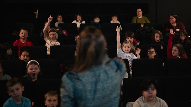 Skupina dětí sedí v kinosále, některé zvedají ruku, zatímco vpředu stojí dospělá osoba zády k nim, pravděpodobně vedoucí nebo lektorka, která s nimi komunikuje.