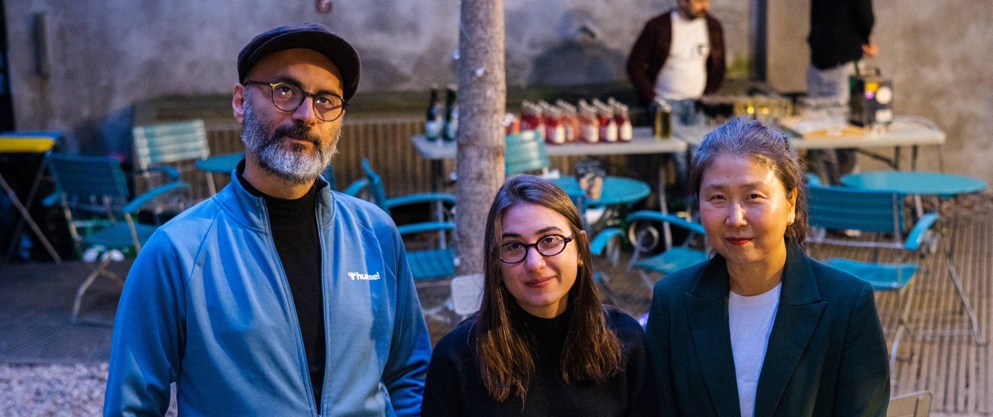 Tři osoby stojí vedle sebe na dvorku s židlemi a stoly.