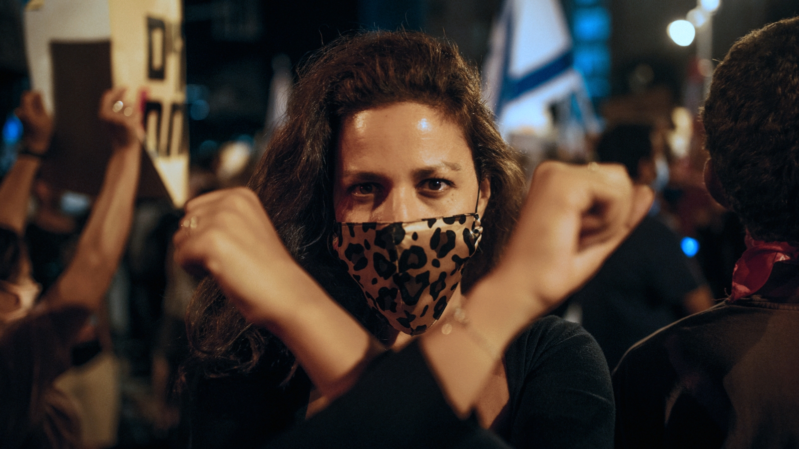 Žena s rouškou s leopardím vzorem stojí v davu lidí na protestu a zvedá ruce.