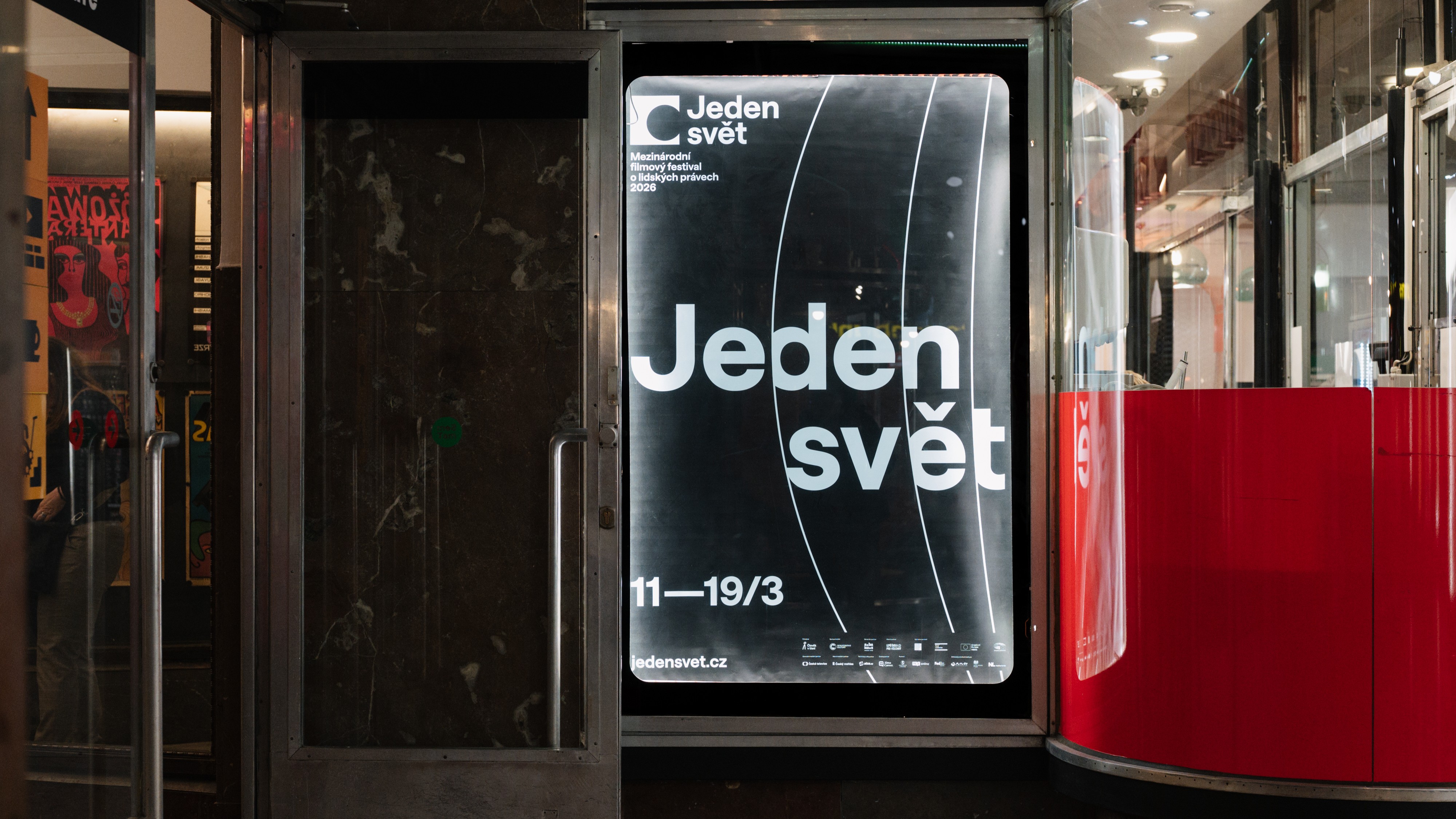 Plakát festivalu Jeden svět ve výloze v pasáži, na kterém je velký bílý nápis „Jeden svět“ a datum konání. Vedle výlohy je vstup do kina Světozor.