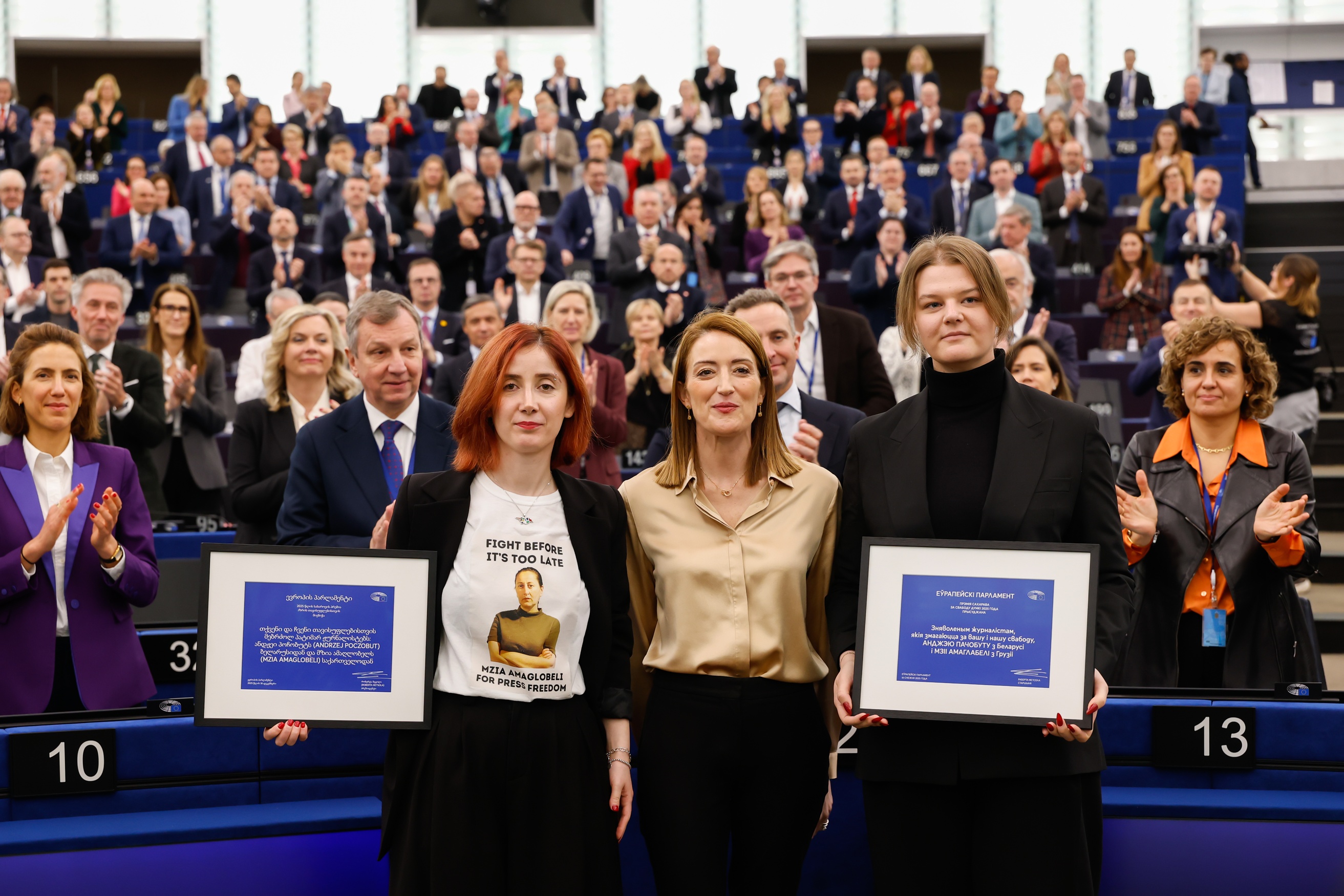 Tři osoby stojí v popředí a drží zarámované dokumenty, zatímco za nimi sedí a stojí zaplněný sál lidí, část publika tleská. Scéna působí jako oficiální událost nebo ceremonie ve velkém konferenčním prostoru.