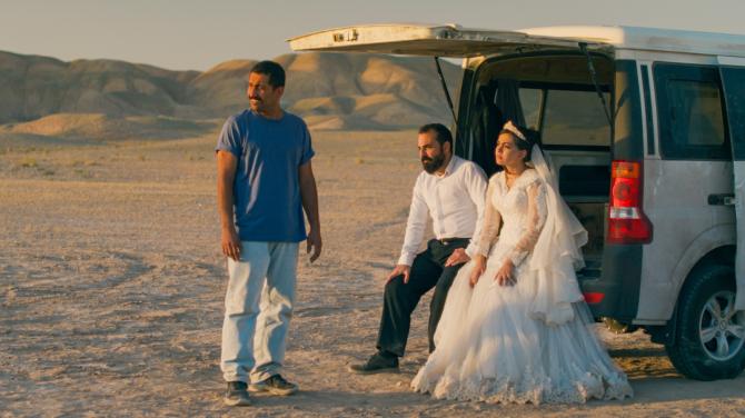 U zaparkované dodávky na prašné pouštní pláni sedí ženich a nevěsta ve svatebním oblečení, oba působí unaveně a zamyšleně. Před nimi stojí muž, všichni se dívají do dálky stejným směrem. V pozadí jsou holé kopce zalité teplým světlem.