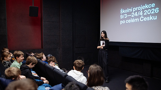 Mladá žena stojí před promítacím plátnem v malém kinosále a mluví ke skupině sedících školních dětí. Na plátně je text „Školní projekce 9/3–24/4 2026 po celém Česku“. Děti sedí v řadách sedaček a poslouchají prezentaci.
