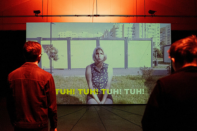 Repeat After Me 2022: Výstava, na které se budete nejdřív smát, než vám dojde, že na ní nic k smíchu není „Dva návštěvníci stojí v temné galerii a sledují velkou projekci videa. Na plátně sedí žena na trávě před panelovými domy a vyslovuje slovo ‚TUH!‘, které se opakuje jako titulky na obrazovce.