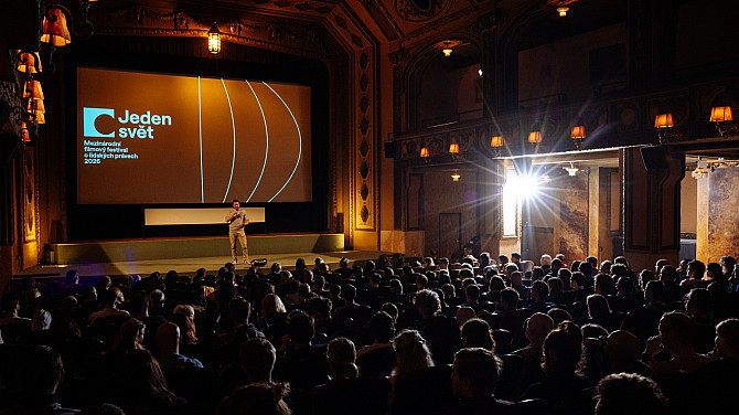 3. den Jednoho světa Moderátor stojí na pódiu v historickém kině a hovoří k plnému sálu diváků. Na velkém plátně za ním je promítán vizuál festivalu Jeden svět – Mezinárodní filmový festival o lidských právech 2026.