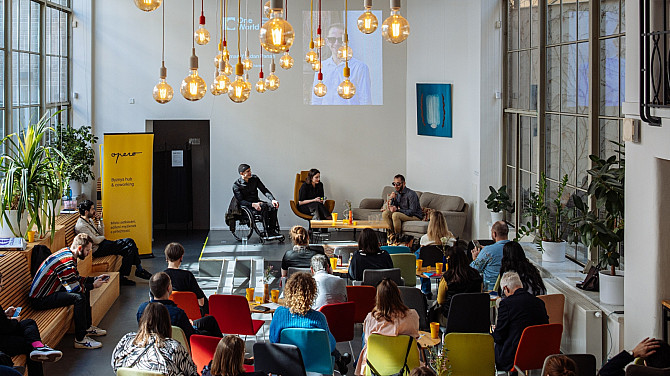 4. den Jednoho světa Panelová diskuse v moderním coworkingovém prostoru, kde tři řečníci sedí na pódiu před publikem; nad nimi visí dekorativní žárovky a za nimi je promítaná prezentace. Posluchači sedí na barevných židlích a sledují debatu.