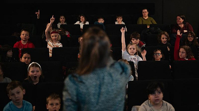 6. den Jednoho světa Skupina dětí sedí v kinosále, některé zvedají ruku, zatímco vpředu stojí dospělá osoba zády k nim, pravděpodobně vedoucí nebo lektorka, která s nimi komunikuje.