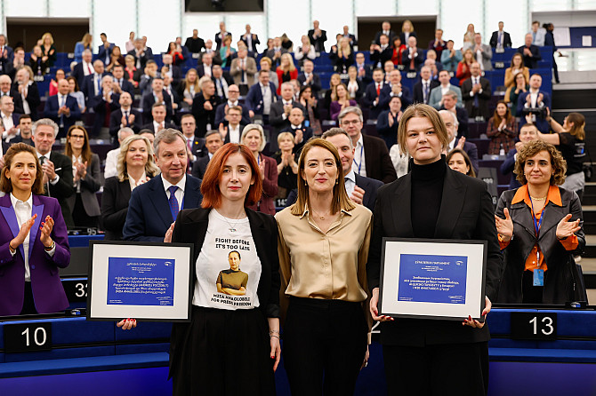 Sacharovovu cenu za rok 2025 získali perzekuovaní novináři z Gruzie a Běloruska Tři osoby stojí v popředí a drží zarámované dokumenty, zatímco za nimi sedí a stojí zaplněný sál lidí, část publika tleská. Scéna působí jako oficiální událost nebo ceremonie ve velkém konferenčním prostoru.