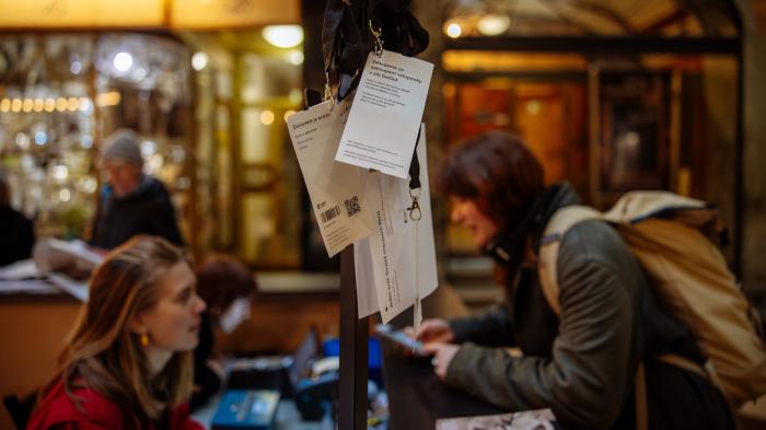 Jak na vstupenky? Na popředí visí několik vstupenek na šňůrce s kolíčky, v pozadí rozostřený infostánek, kde si žena s batohem kupuje lístek a naproti ní sedí žena za pultem.