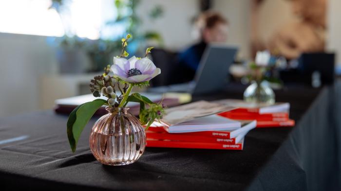 Na stole s černým ubrusem stojí váza s kyticí a štos katalogů.