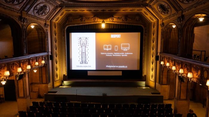 Rozsvícený kinosál před projekcí. Na plátně běží předfilmové spoty.