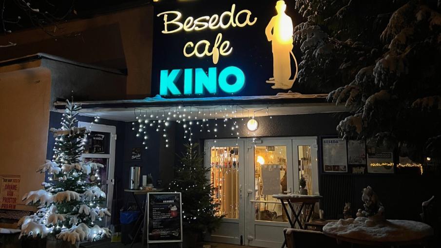 Vstupní dveře do budovy kina, vlevo ozdobený vánoční stromeček, vpravo posezení. Nad vstupem svítící nápis Beseda cafe kino.