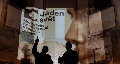 Dvě postavy stojí ve tmě a kontrastují s projekcí festivalového vizuálu. 