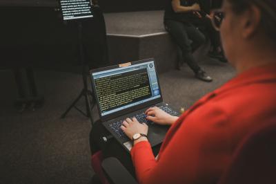 Osoba zapisuje na notebooku simultánní přepis pro neslyšící účastníky debaty. V pozadí sedí lidé a na stojanu je vidět další monitor s promítaným přepisem.