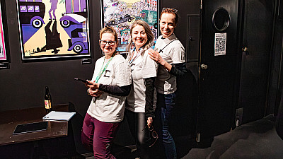 Tři mladé ženy stojí za sebou a se usmívají se. Na sobě mají trička s logem filmového festivalu Jeden svět a na krku pověšené cedulky Pořadatel.