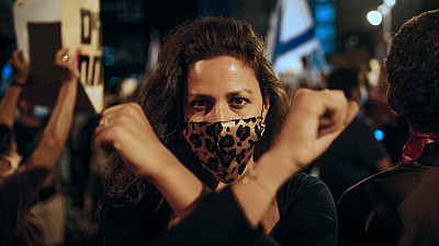 Žena s rouškou s leopardím vzorem stojí v davu lidí na protestu a zvedá ruce.