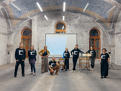 Skupina několika lidí ve stejných festivalových mikinách a tričkách stojí v prostorné místnosti s klenutým stropem, za nimi je projekční plátno a okna.